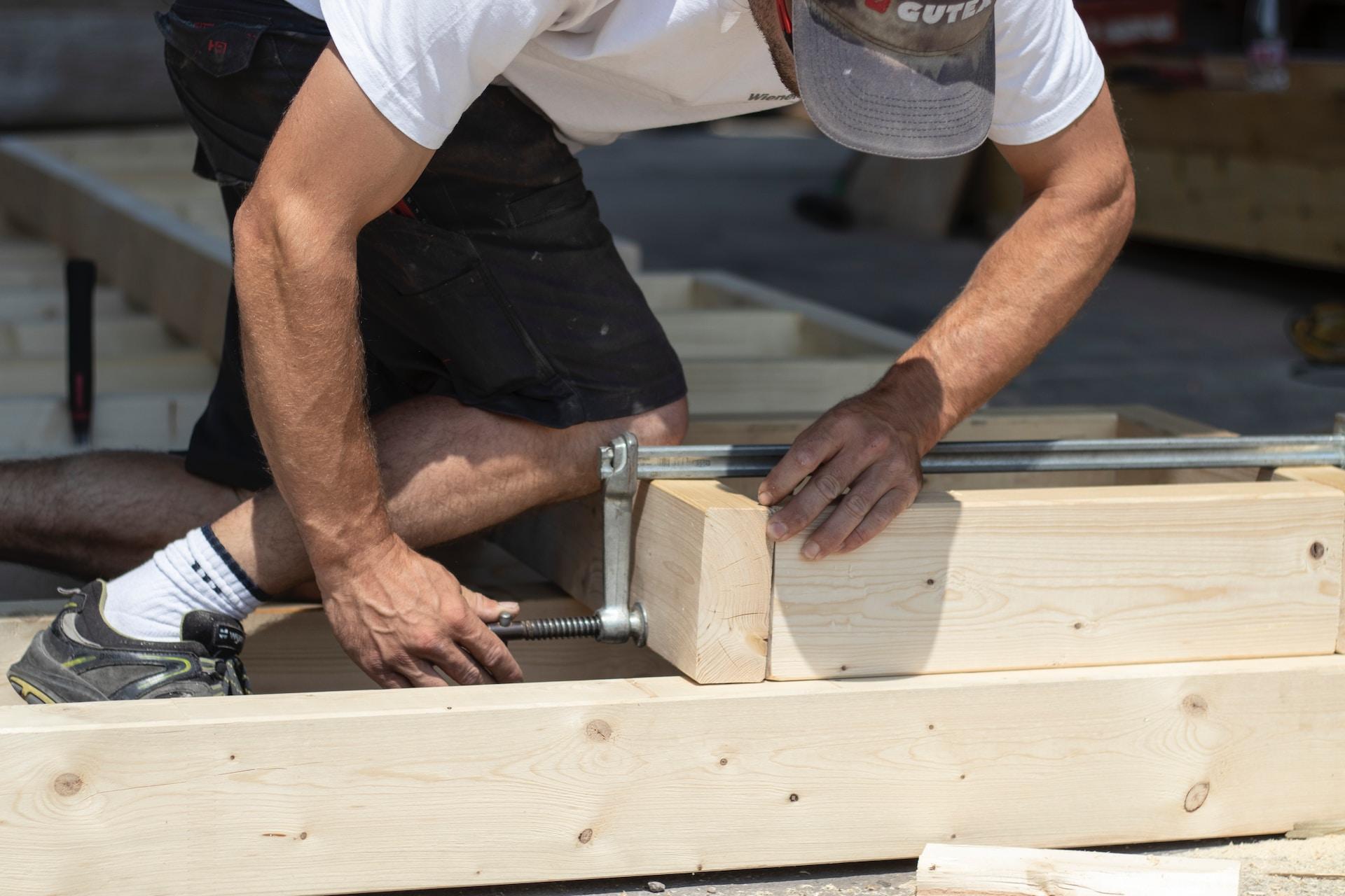 Ein Zimmerer kniet auf einem Holzbalken und zieht eine Schraubzwinge an